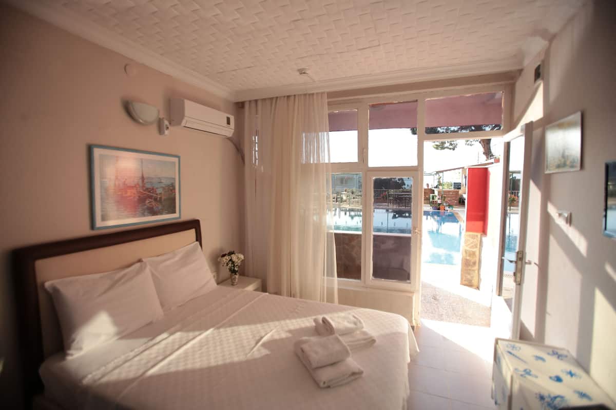 Une chambre d’hôtel avec un lit, une fenêtre donnant sur une piscine, un climatiseur fixé au mur et un tableau encadré.