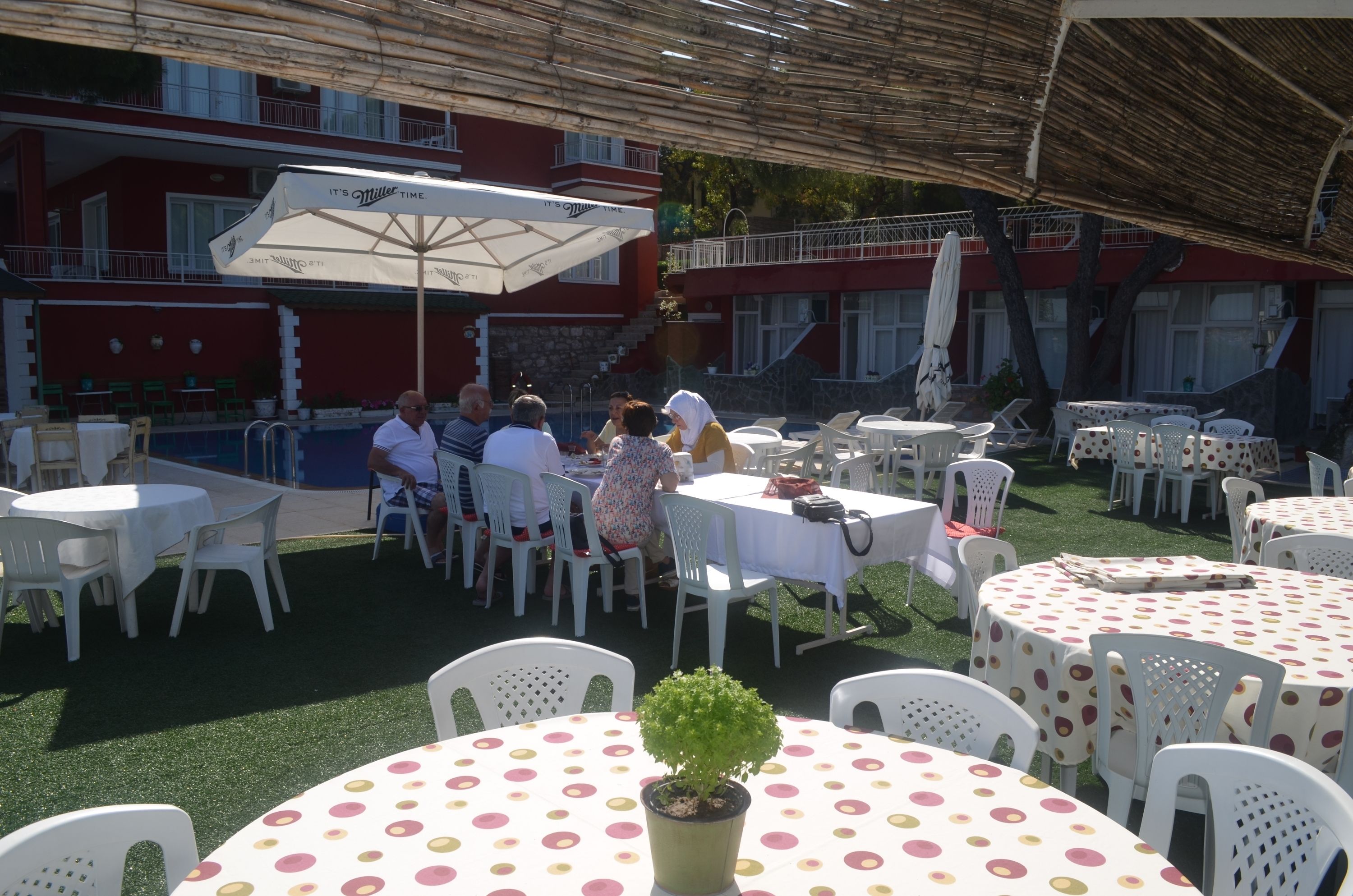 An outdoor dining area with round tables and chairs, a pool, and a building with balconies in the background.