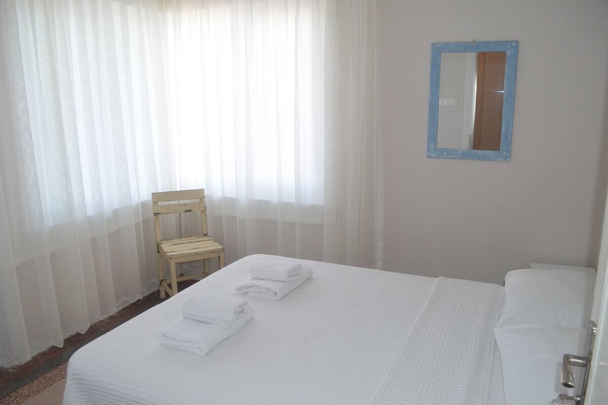 Une chambre d’hôtel avec un lit, une chaise, un miroir et une fenêtre avec des rideaux légers.