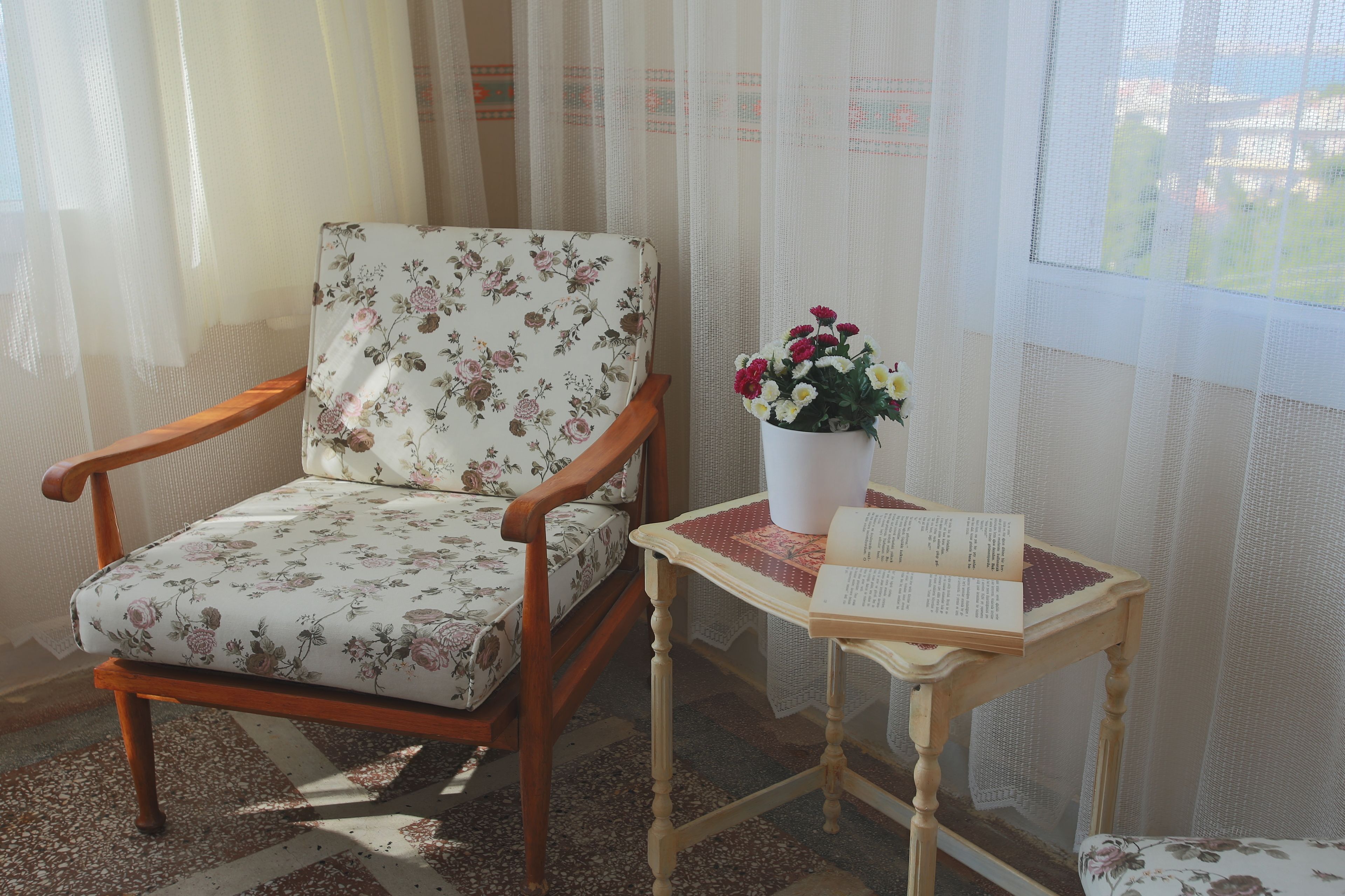 A room with a wooden chair with floral upholstery, a small wooden table with a book and a potted plant.