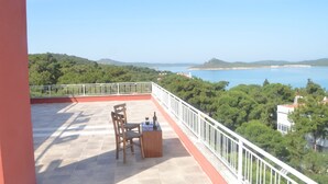 A terrace with a table and chairs, overlooking a coastal landscape with trees and a view of the sea.