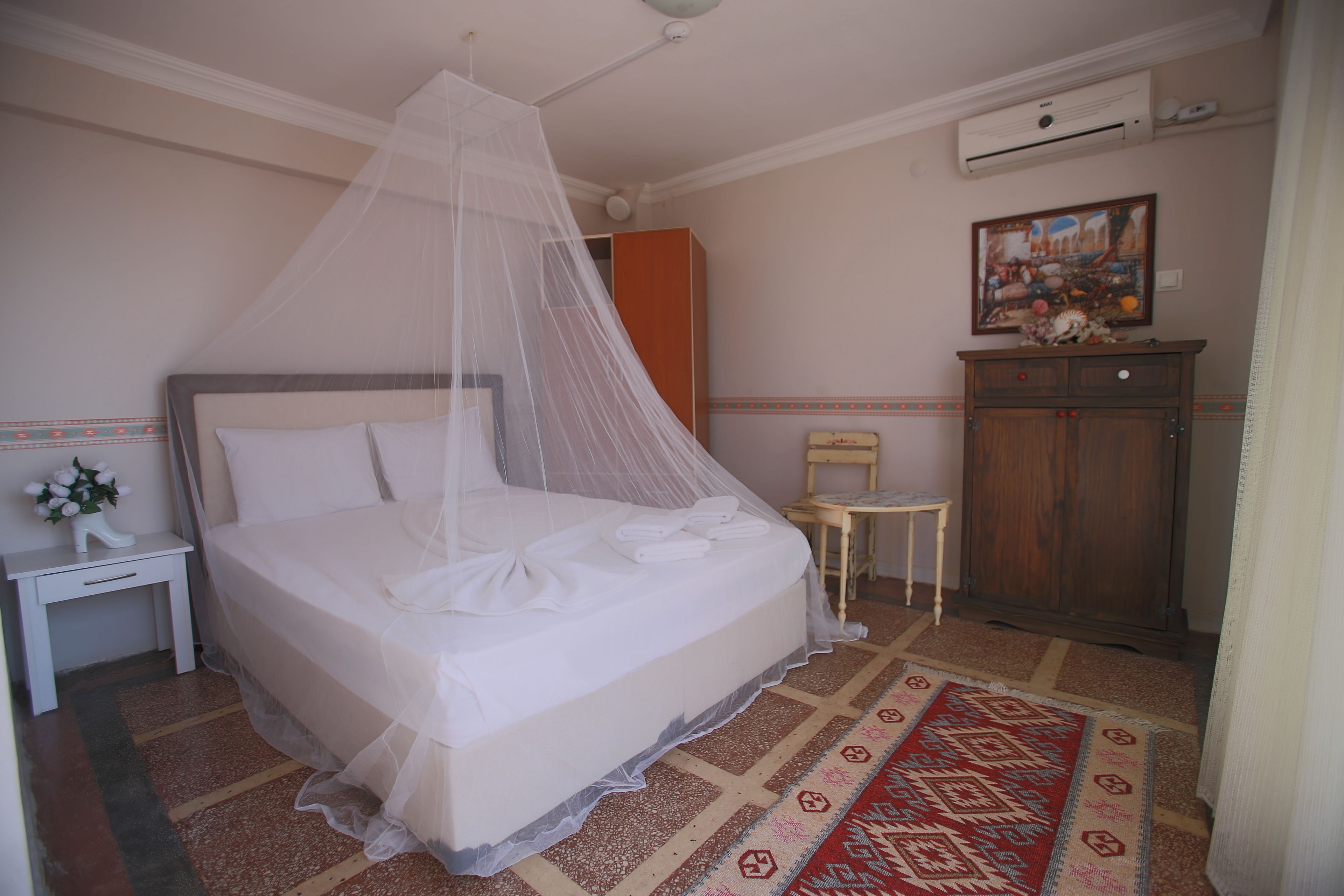 A neatly made bed with a canopy, a small bedside table with a lamp, a wooden cabinet, and a patterned rug.