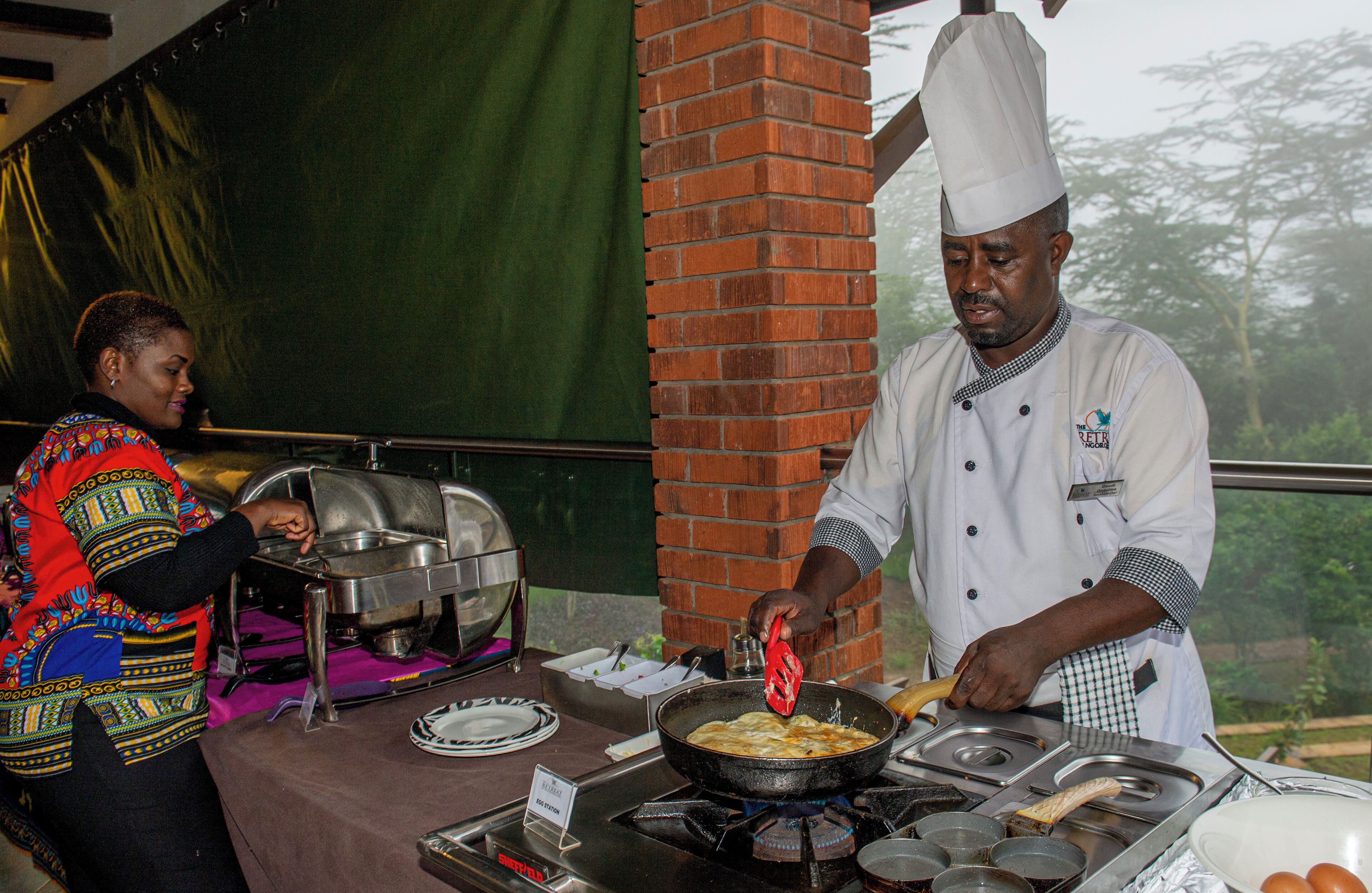 A chef cooking outdoors with a brick wall and a green tarp in the background.
