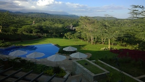 A pool area with umbrellas, a lawn, and a view of a forested landscape. - The Retreat at Ngorongoro (Karatu)