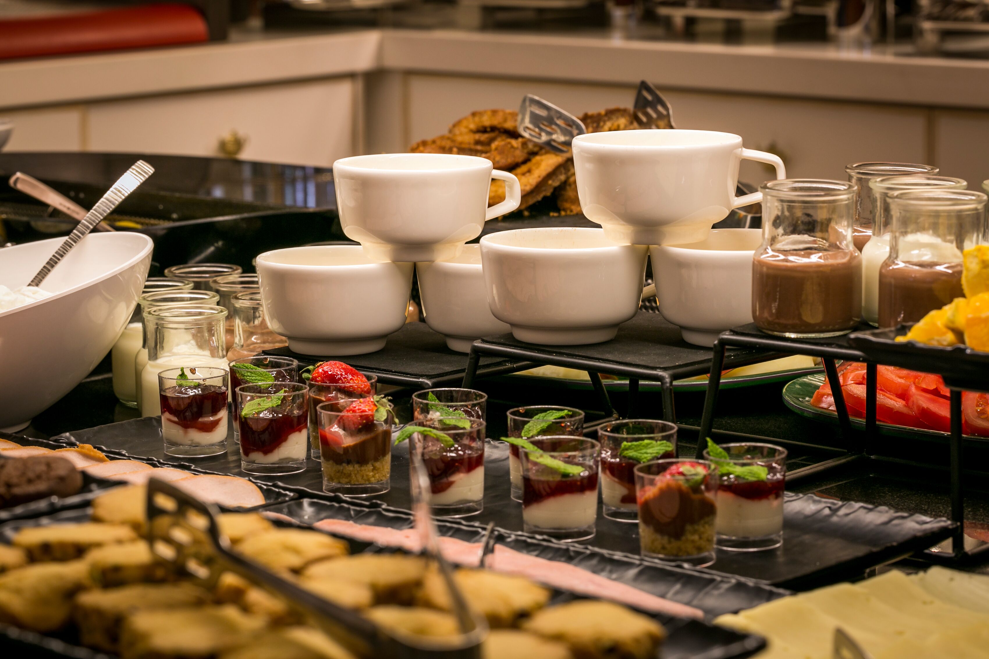 Ein Buffet mit verschiedenen Desserts und Getränken, darunter mehrschichtige Desserts in Gläsern, eine Schüssel mit Keksen sowie mehrere weiße Tassen und Gläser.