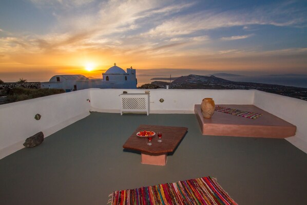 A rooftop terrace with a sunset view, a white building, and a table with a bowl of fruit and two glasses. - Skyline Villa (Santorini)