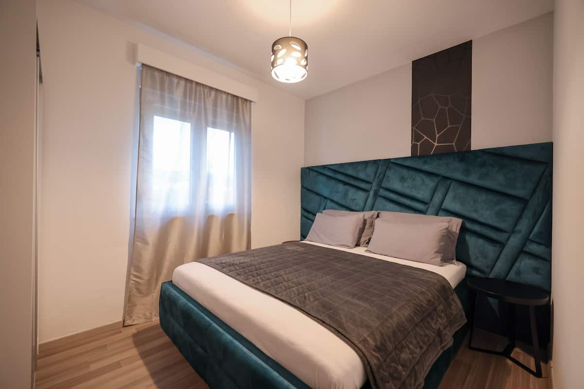 Un dormitorio moderno con una cama grande, cabecero, un taburete pequeño y una ventana con cortinas.