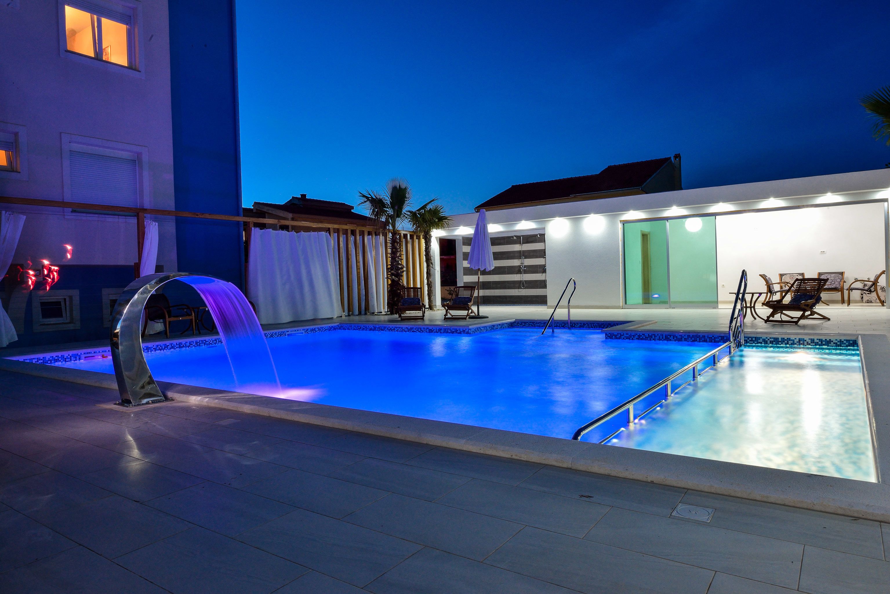 Una piscina con fuente acuática, rodeada por un patio cubierto con asientos y una puerta que da a un espacio techado.