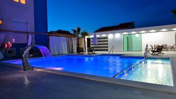 Una piscina con fuente acuática, rodeada por un patio cubierto con asientos y una puerta que da a un espacio techado.