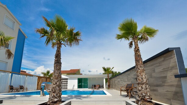 Een modern zwembadgedeelte met palmtoppen, een helderblauwe lucht en een gebouw met een blauwe gevel en een houten terras.