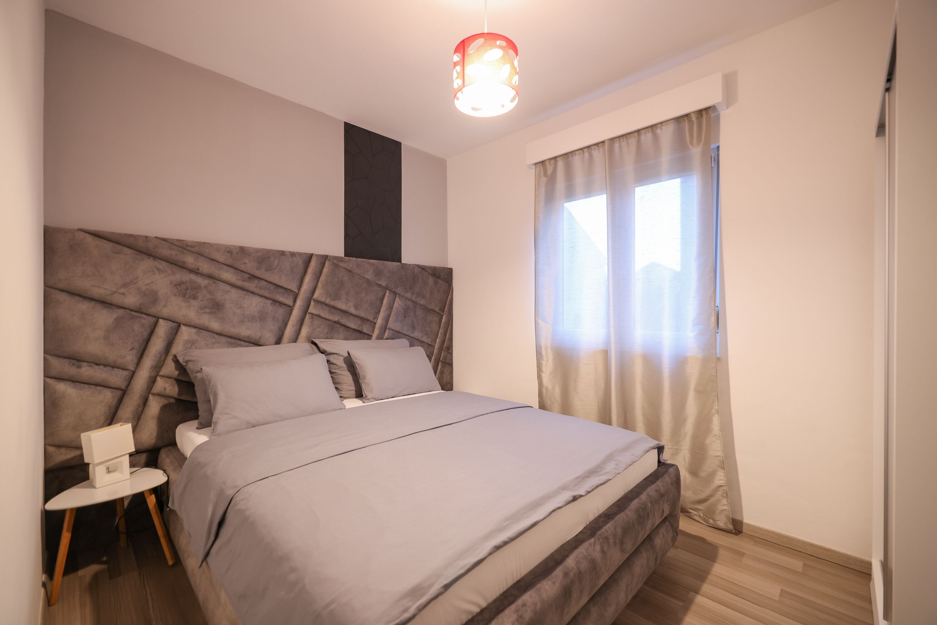 A modern bedroom with a large bed, a nightstand, a wall-mounted light fixture, and a window with curtains.