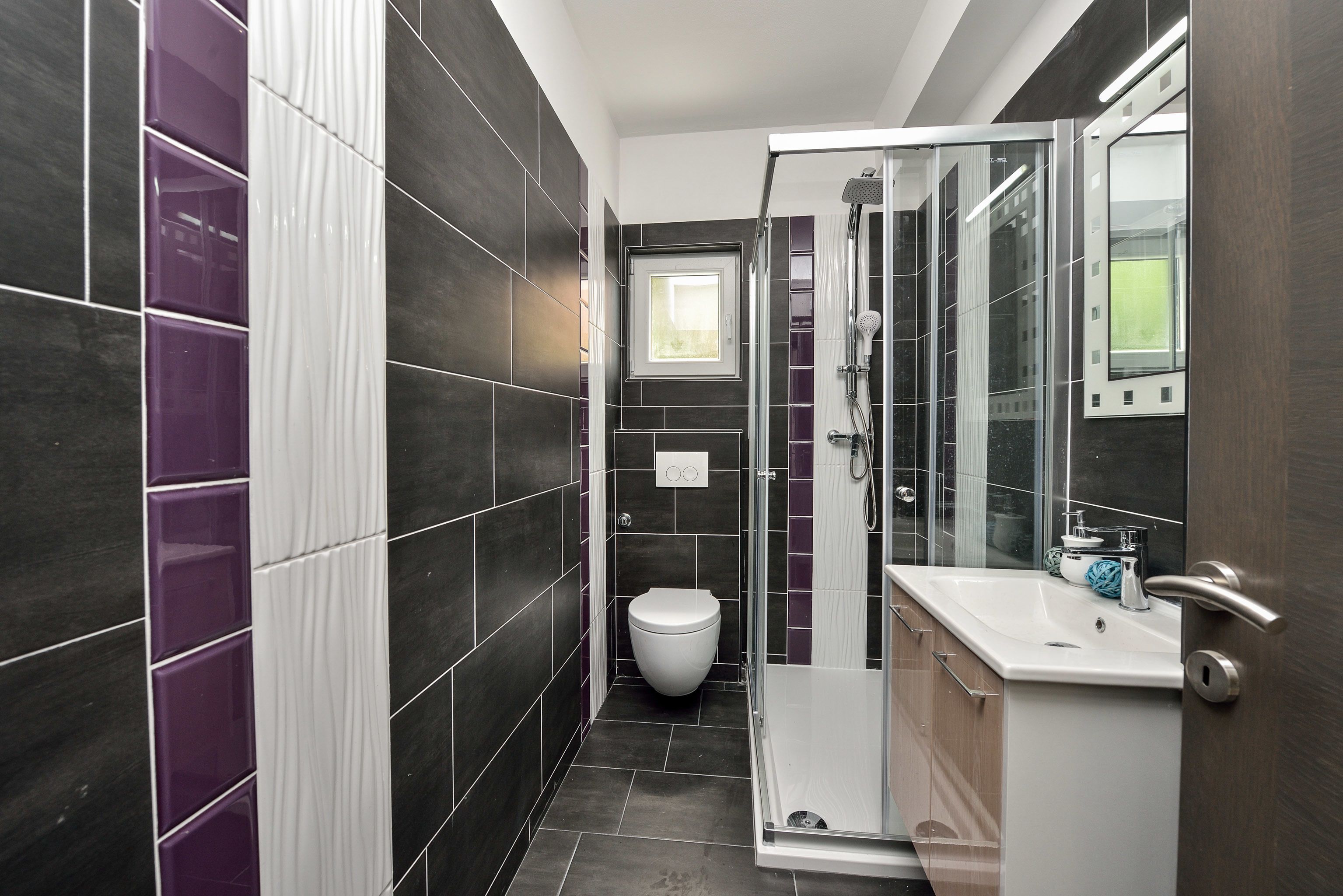 A modern bathroom with dark tiles, a white toilet, a glass shower enclosure, and a sink with a mirror.