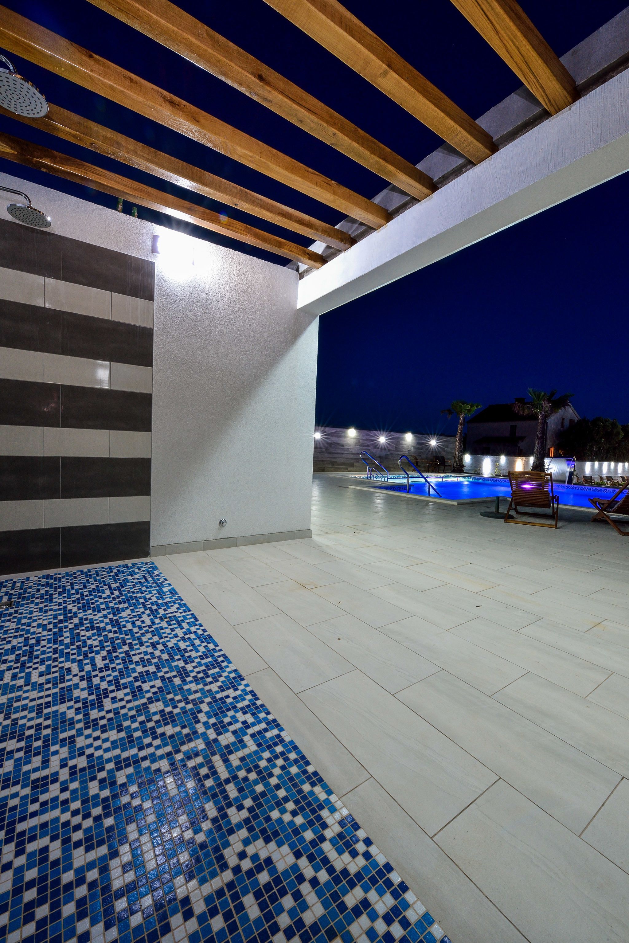 An outdoor swimming pool area with tiled flooring, wooden beams, and a tiled wall.