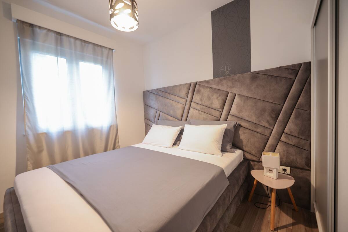 Un dormitorio moderno con una cama grande, cabecera, mesita de noche y ventana con cortinas.