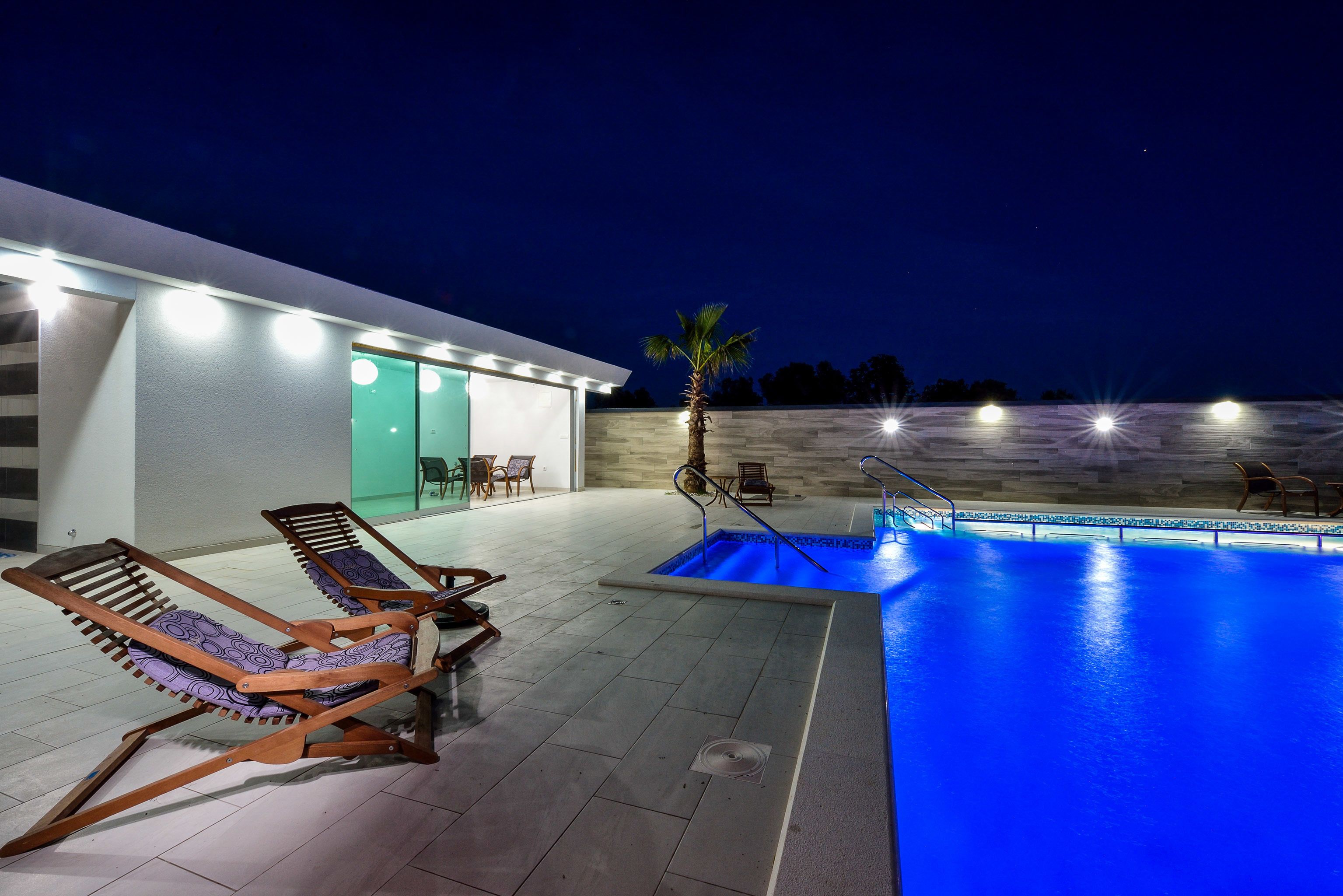 A modern outdoor pool area with lounge chairs, a palm tree, and a building with illuminated windows.