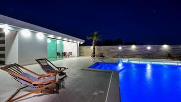 A modern outdoor pool area with lounge chairs, a palm tree, and a building with illuminated windows.