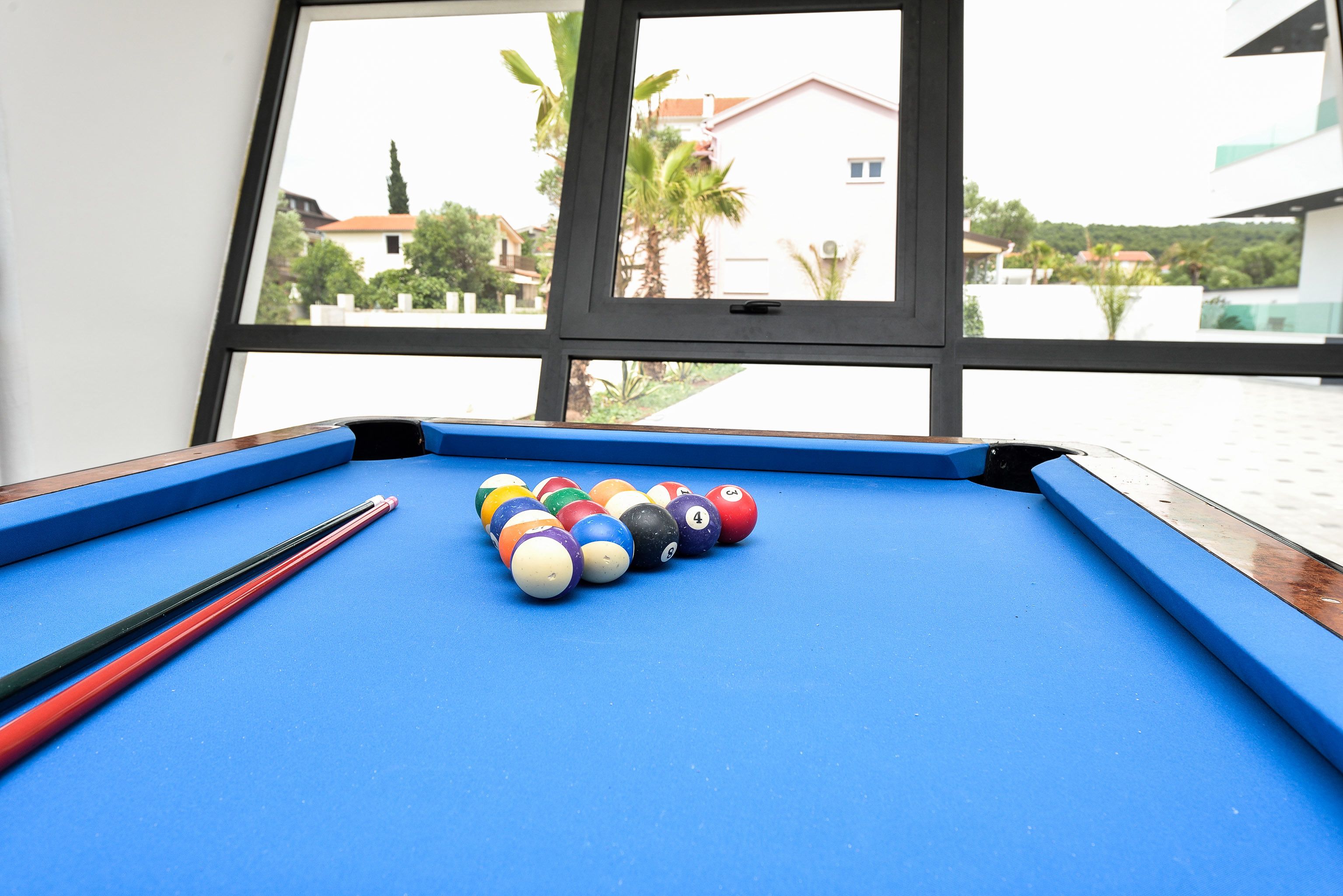 A pool table with billiard balls and cues, positioned near a window with a view of buildings and palm trees.
