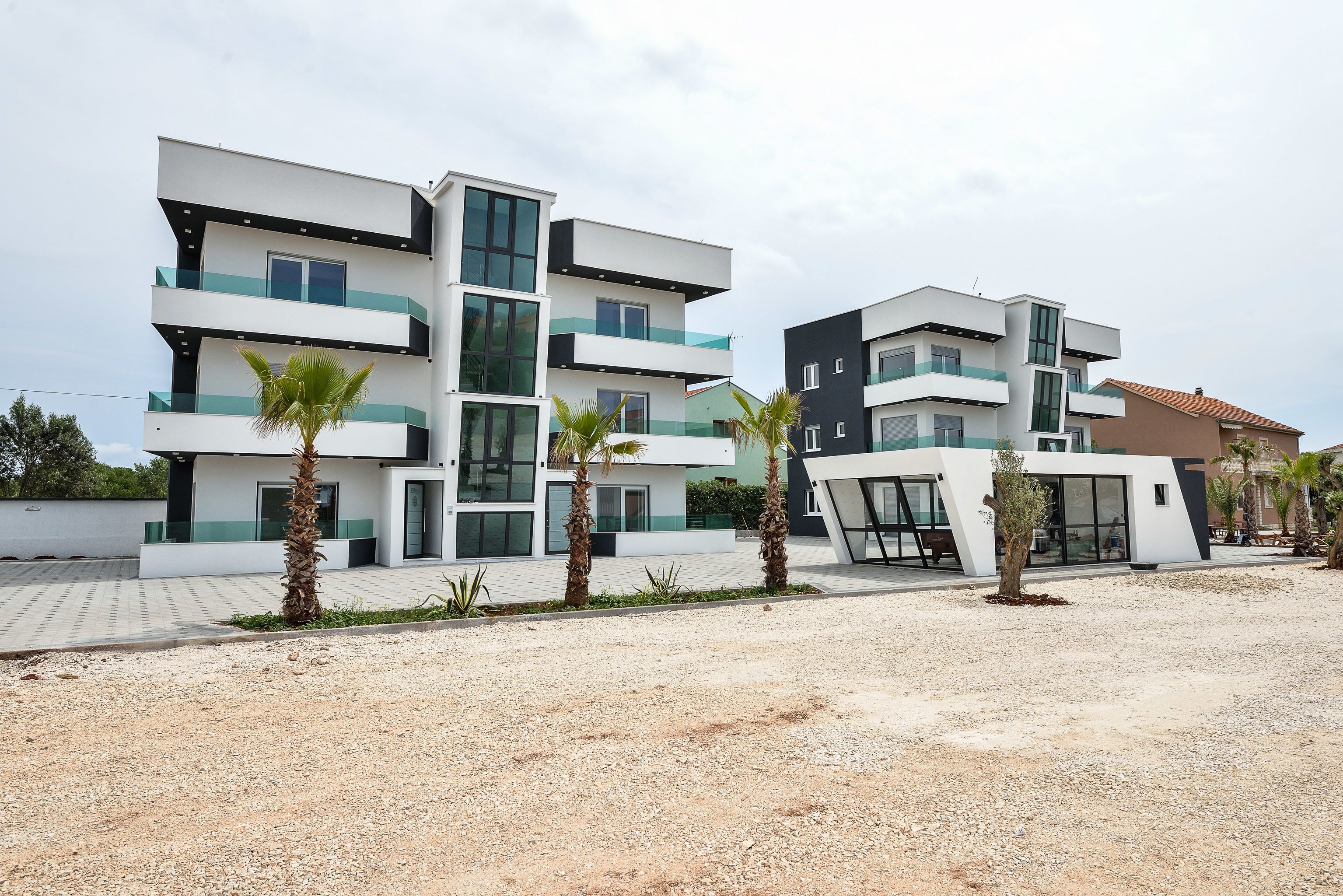 A modern multi-story residential building with balconies, palm trees, and a gravel driveway.
