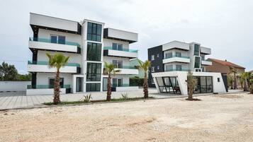 A modern multi-story residential building with balconies, palm trees, and a gravel driveway.