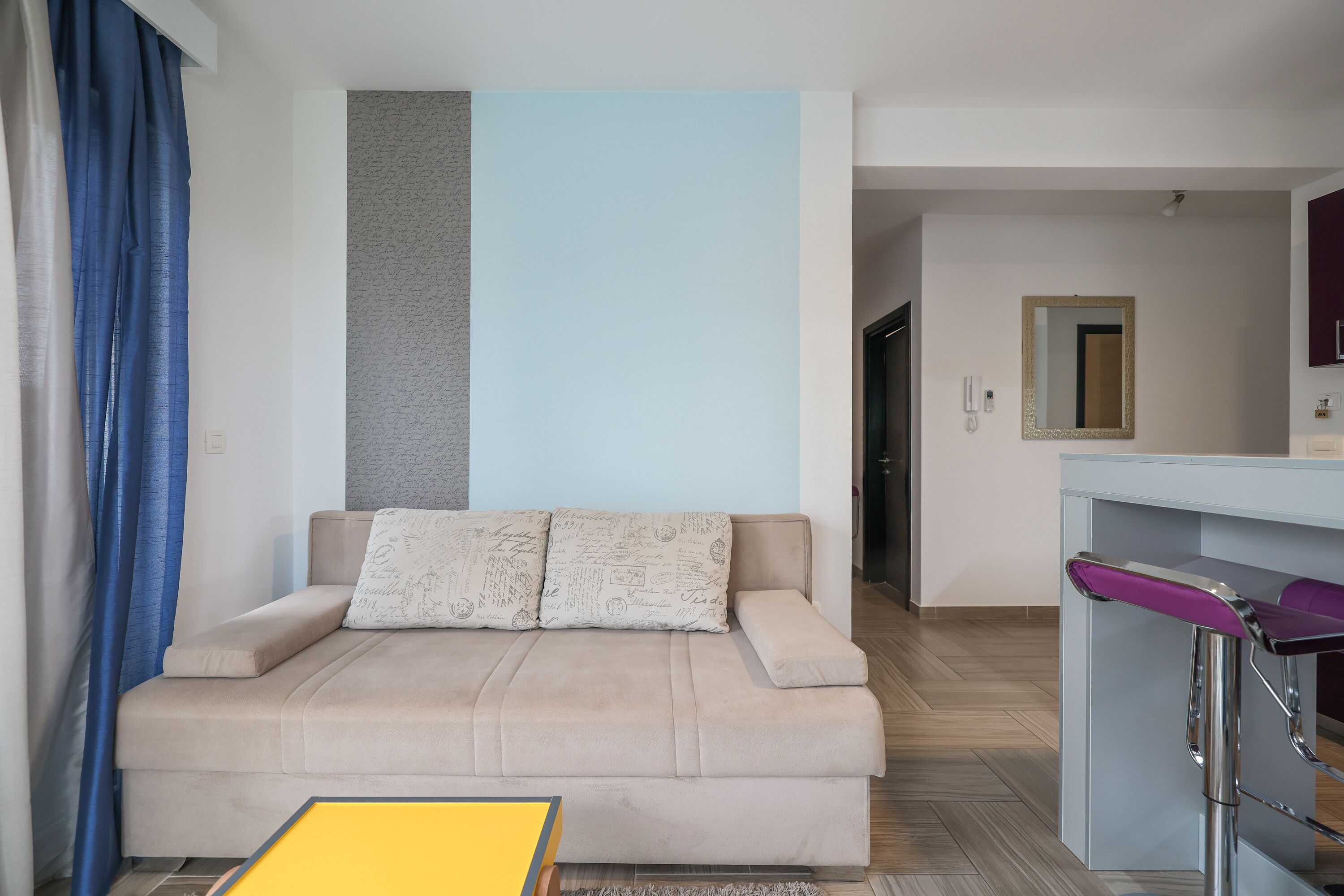 A modern living room with a beige sofa, a small yellow table, and a purple bar stool.