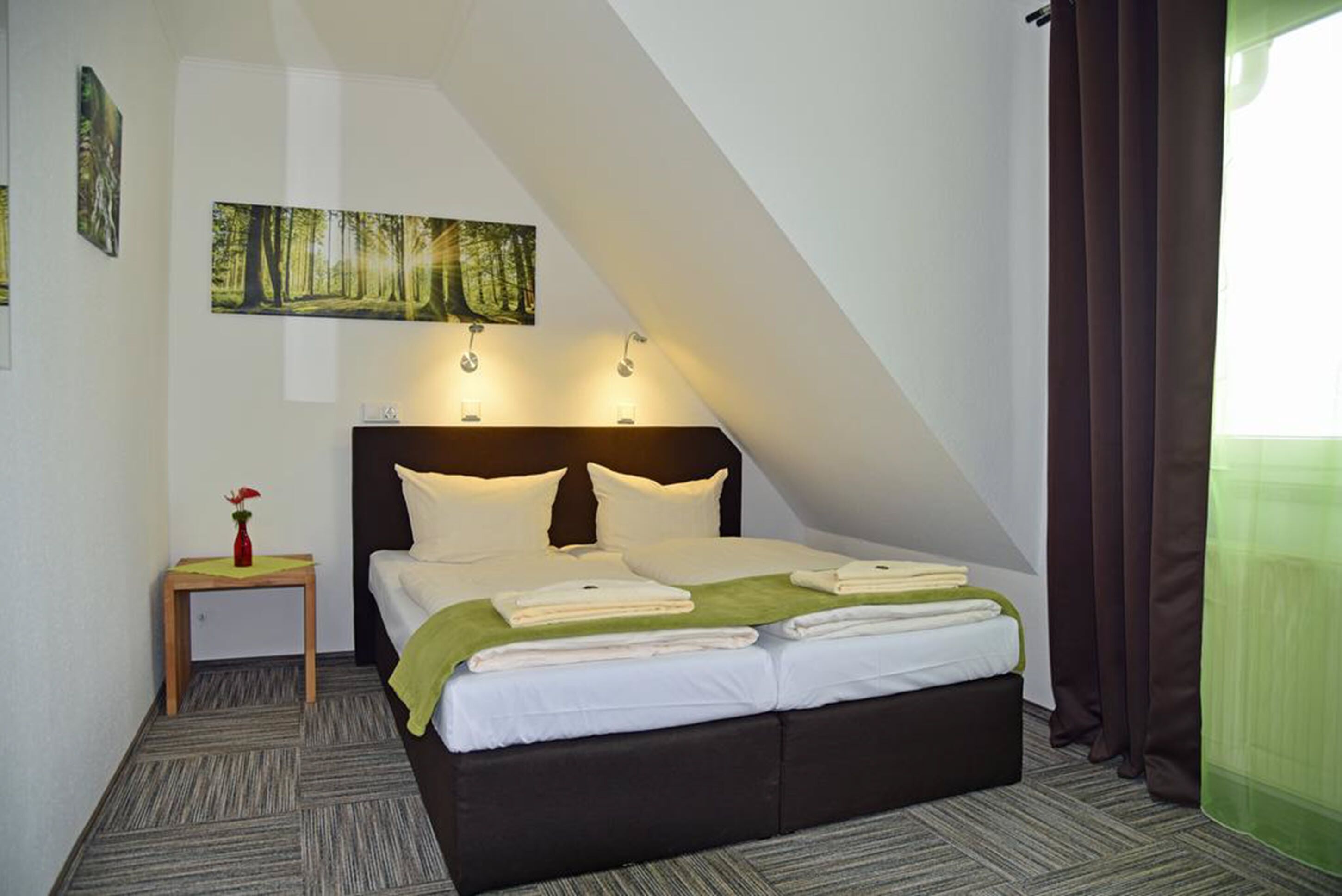 A hotel room with a large bed, a small bedside table with a vase, and a painting on the wall.