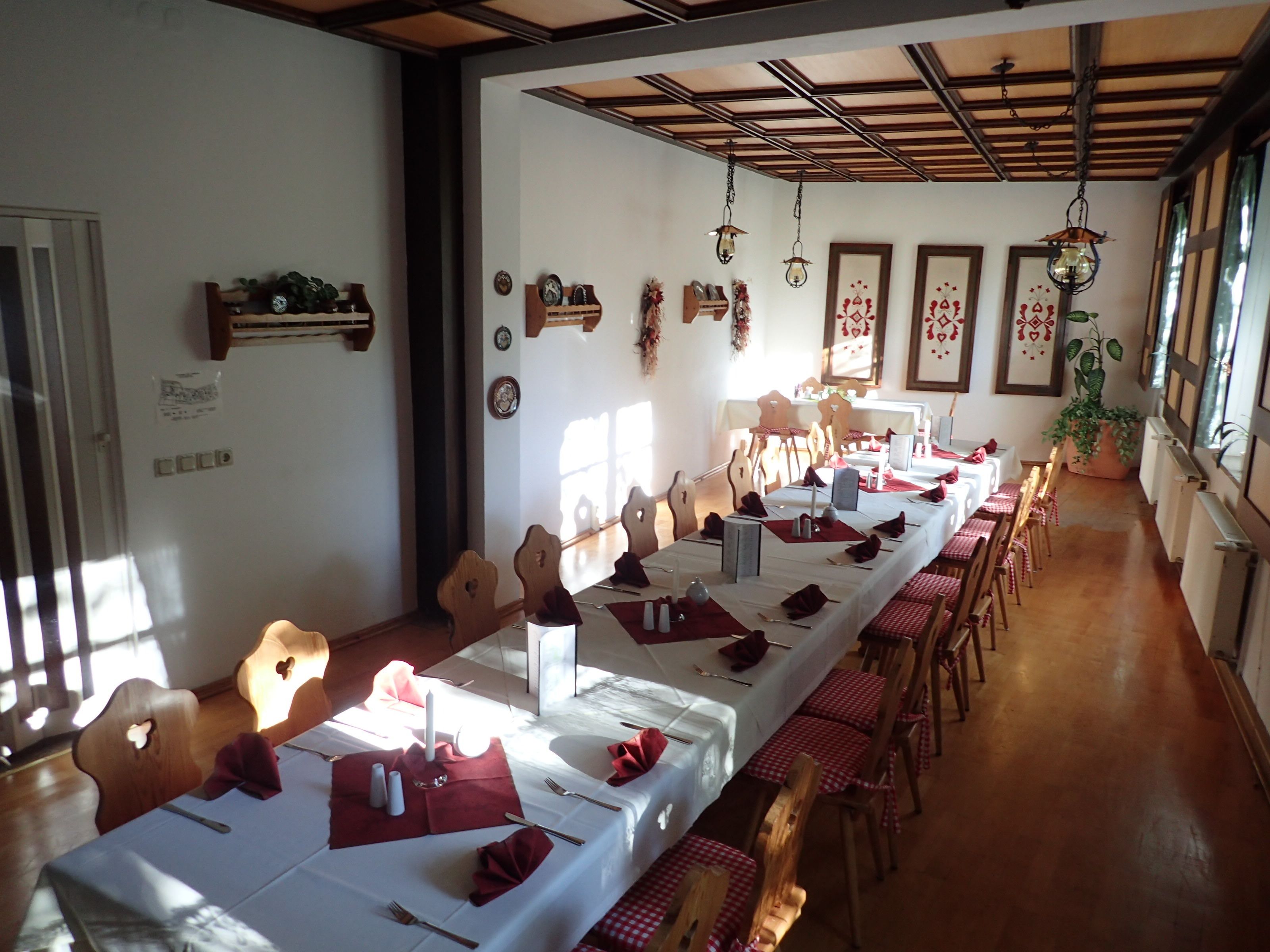 Uma sala de jantar com mesas longas preparadas para uma refeição, cadeiras de madeira e luminárias pendentes.