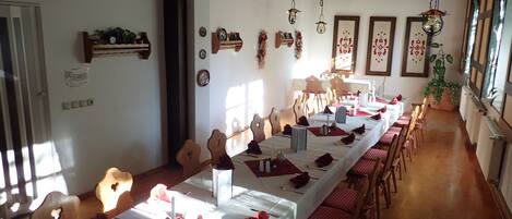 Uma sala de jantar com mesas longas preparadas para uma refeição, cadeiras de madeira e luminárias pendentes.