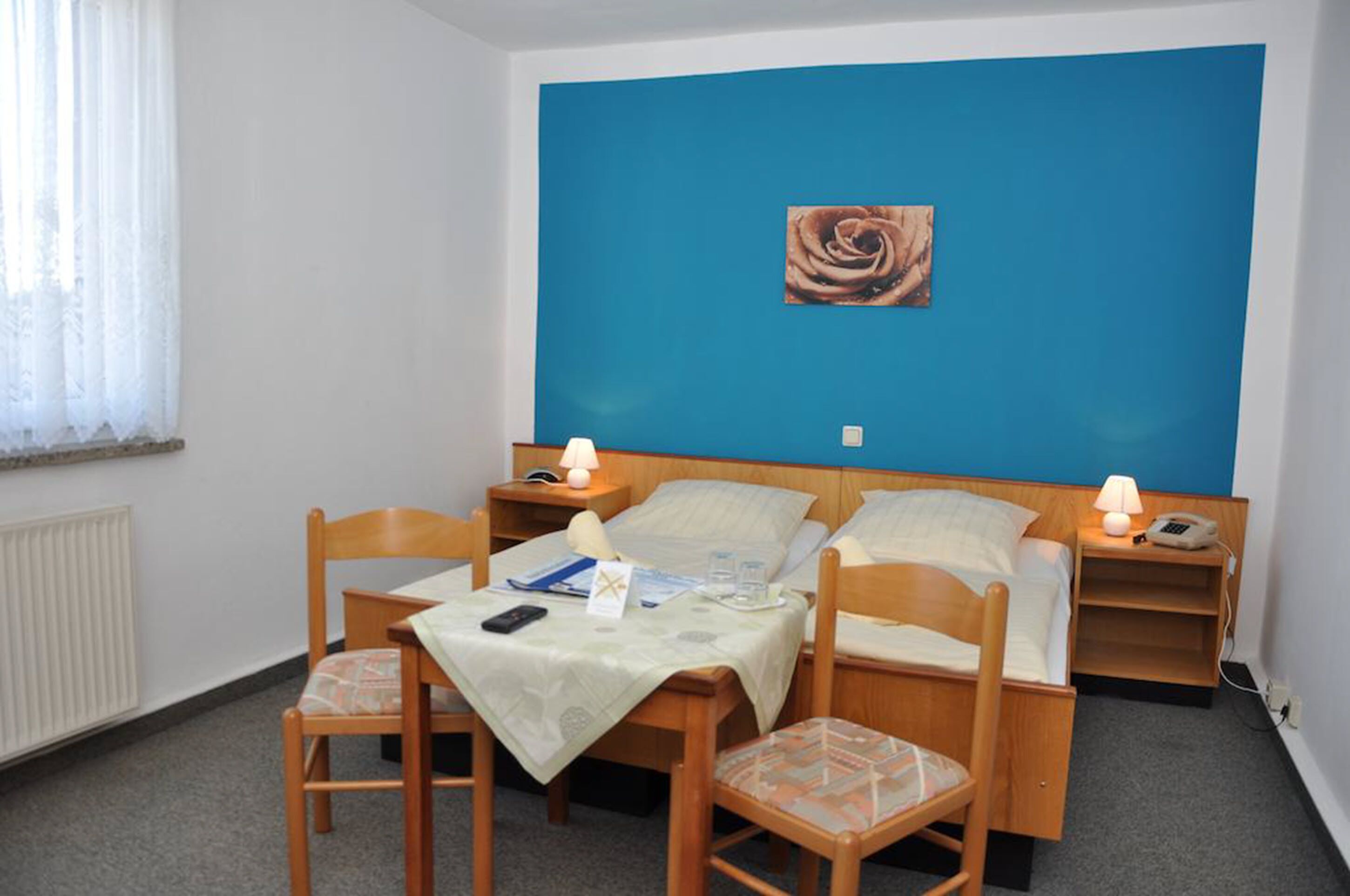 A single bedroom with a blue accent wall, a bed with two pillows, a bedside table with a lamp, a chair, and a small table with a phone and a book.