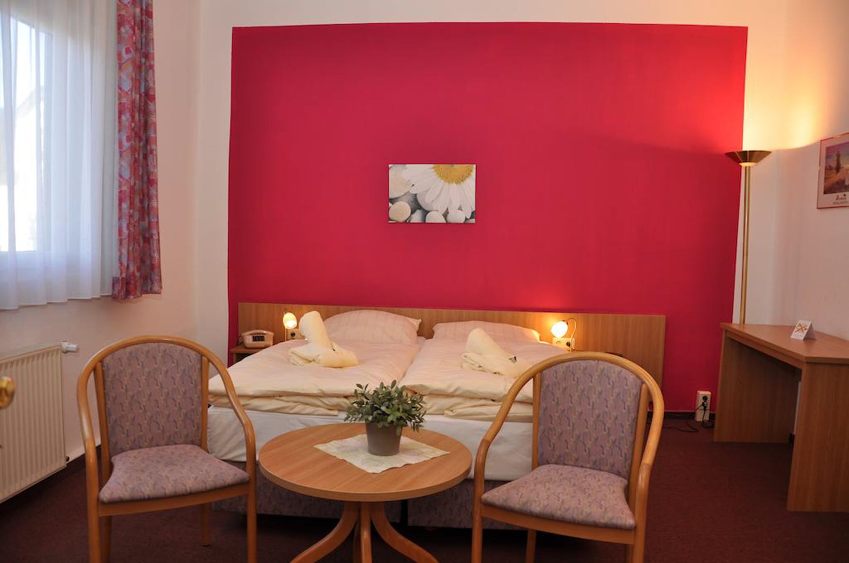 A hotel room with a red accent wall, a bed with white linens, two upholstered chairs, a small round table with a plant, and a bedside lamp.