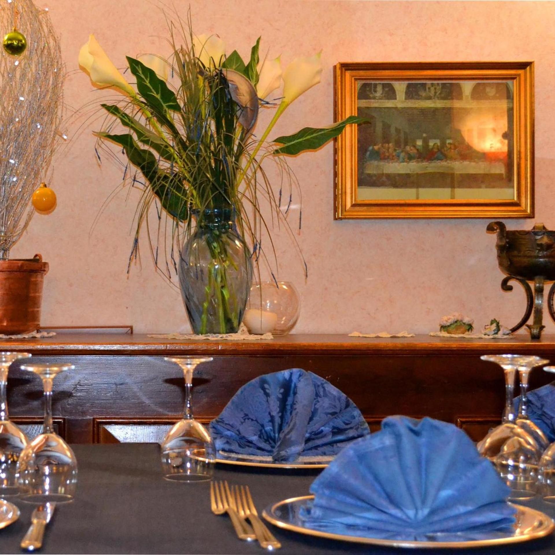 Ein Esstisch ist gedeckt, mit blauen Servietten, einer Vase mit weißen Blumen und einem gerahmten Bild an der Wand.