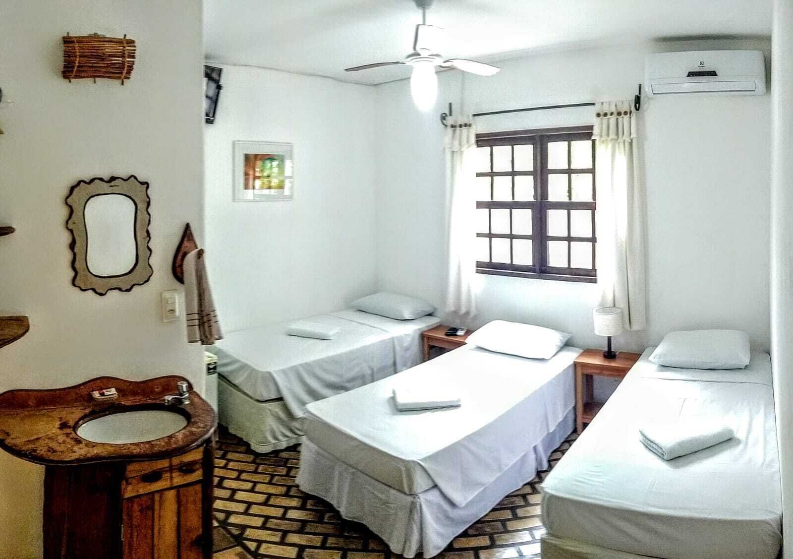 Una habitación pequeña y limpia con dos camas individuales, un inodoro de madera, una ventana con cortinas, un ventilador de techo y un aire acondicionado de pared.