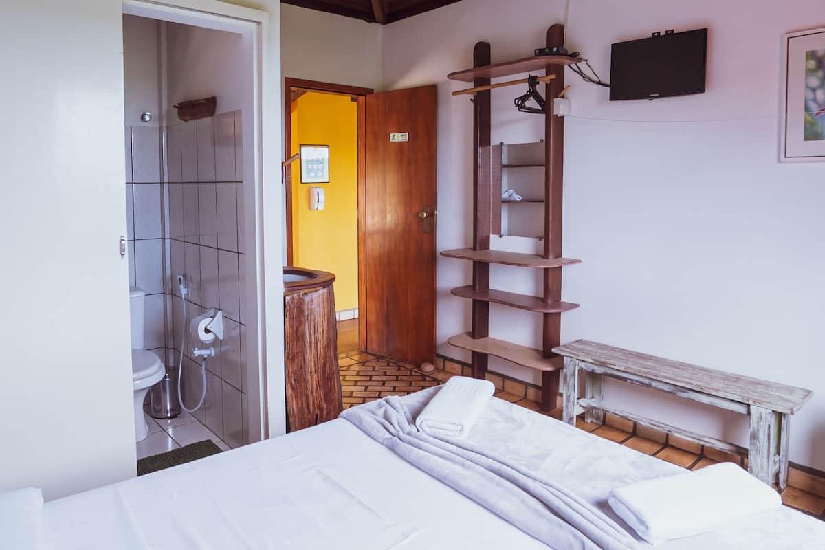 A bedroom with a wooden ceiling, a bed with white bedding, a wooden ladder, a small table, and a bathroom with a shower and toilet.
