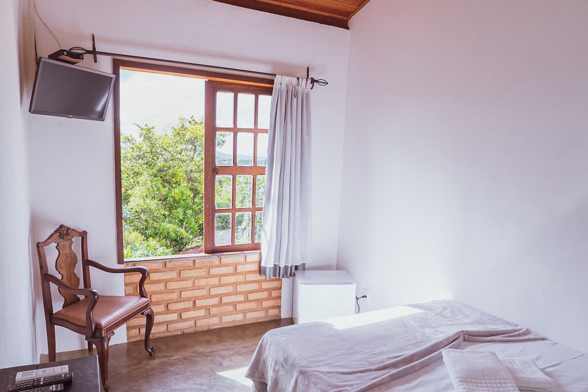 A room with a bed, a chair, a table, a ceiling fan, a window with curtains, and a TV mounted on the wall.