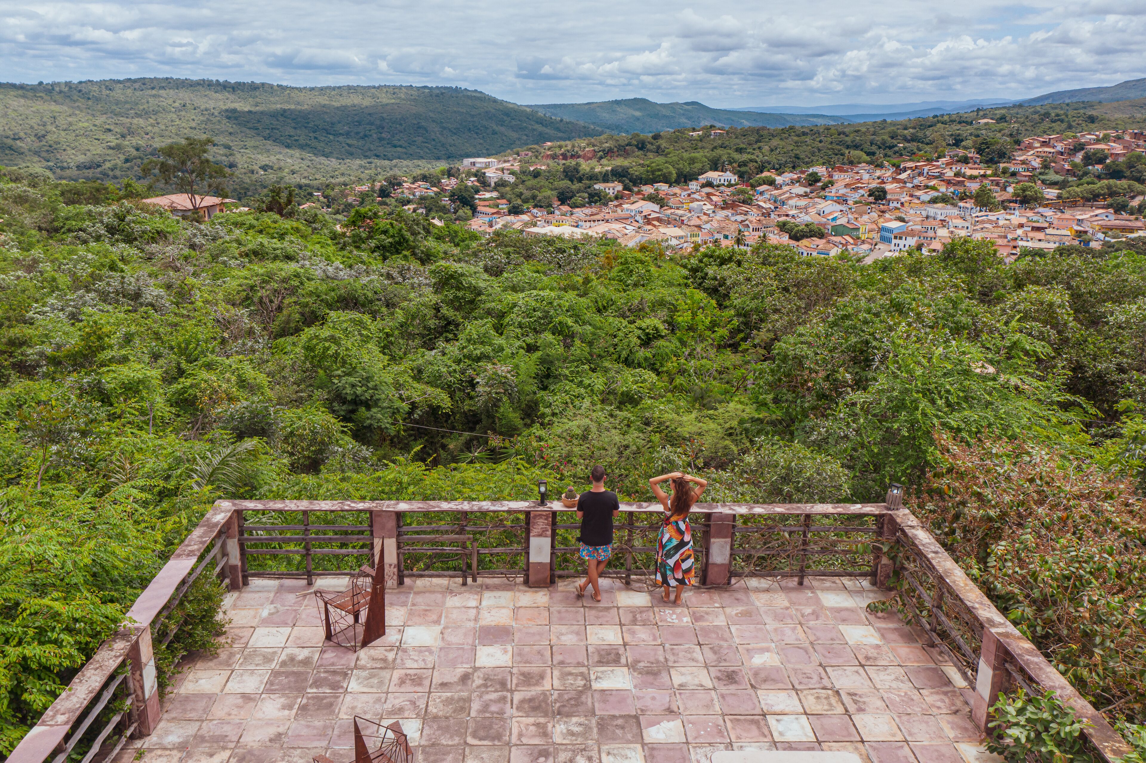 A paved viewing platform with a wooden railing overlooking a town surrounded by greenery.