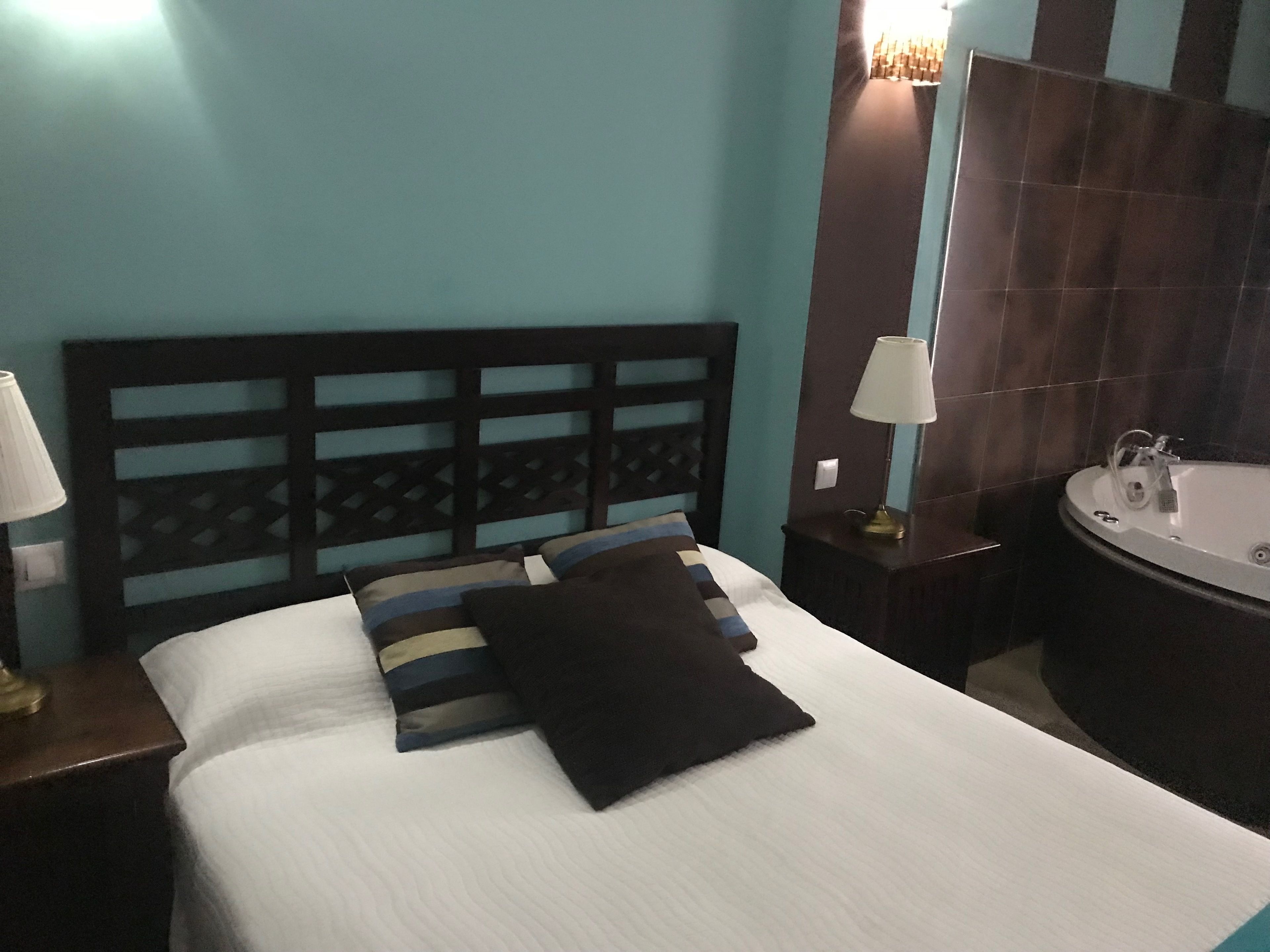 Uma cama bem arrumada com cabeceira, duas mesas de cabeceira com abajures, e um banheiro com banheira e pia.