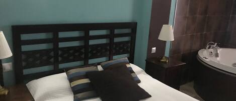 Uma cama bem arrumada com cabeceira, duas mesas de cabeceira com abajures, e um banheiro com banheira e pia.