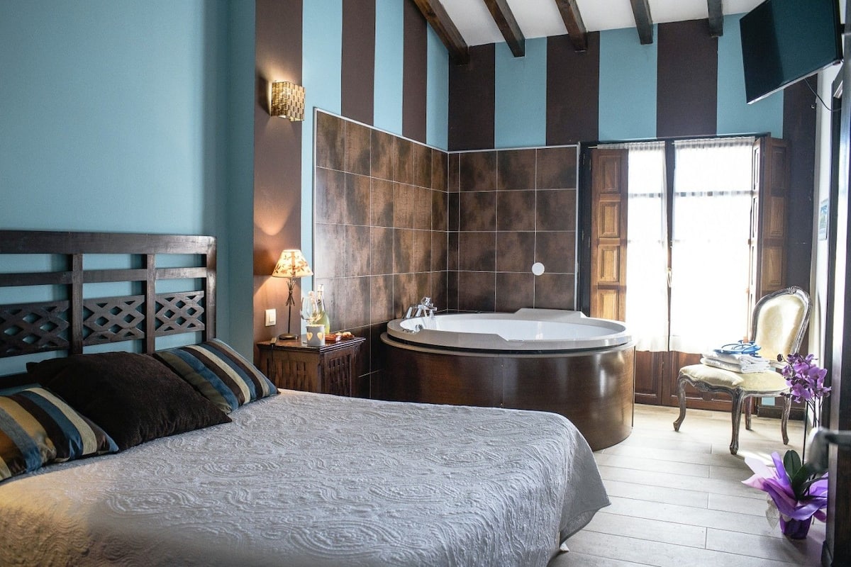 Un dormitorio con una cama grande, una zona de estar con una silla, una bañera y una ventana con cortinas.