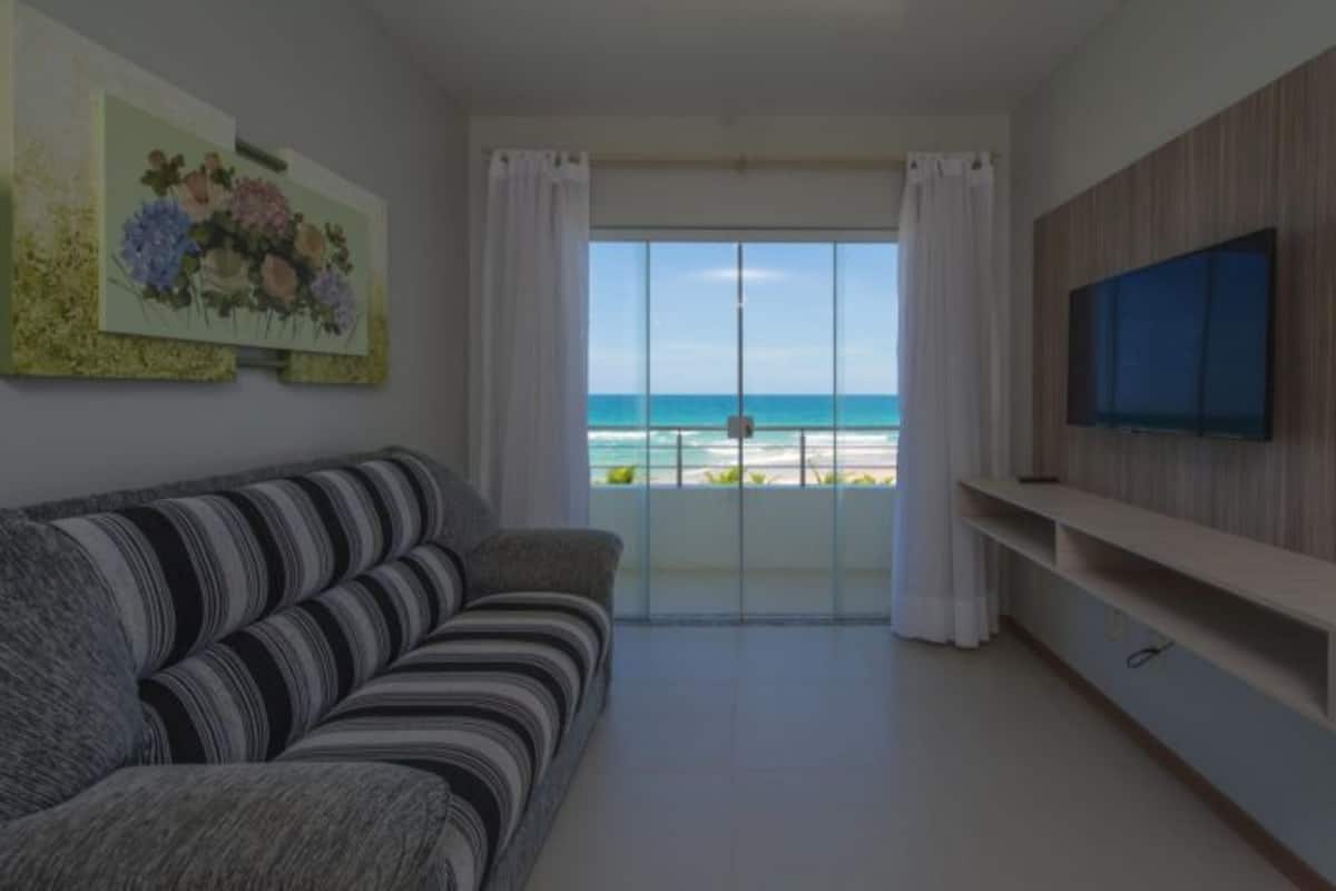 A living room with a sofa, a TV, and a balcony with a view of the beach.