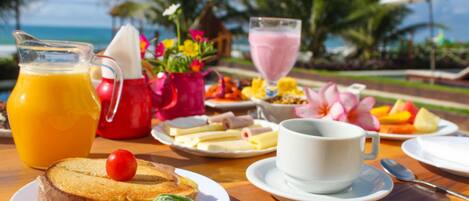Una mesa de desayuno con una jarra de jugo de naranja, una tetera roja, una taza blanca, platos con comida y vistas al océano.