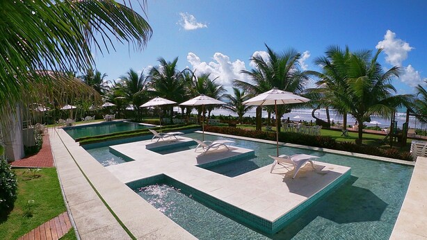 A rectangular swimming pool with clear water, surrounded by palm trees and lounge chairs with umbrellas. - Hotel Pousada Butias   (Ipojuca)