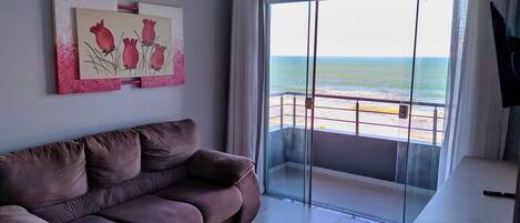 A living room with a brown sofa, a painting of flowers, and a view of the beach through a glass door.