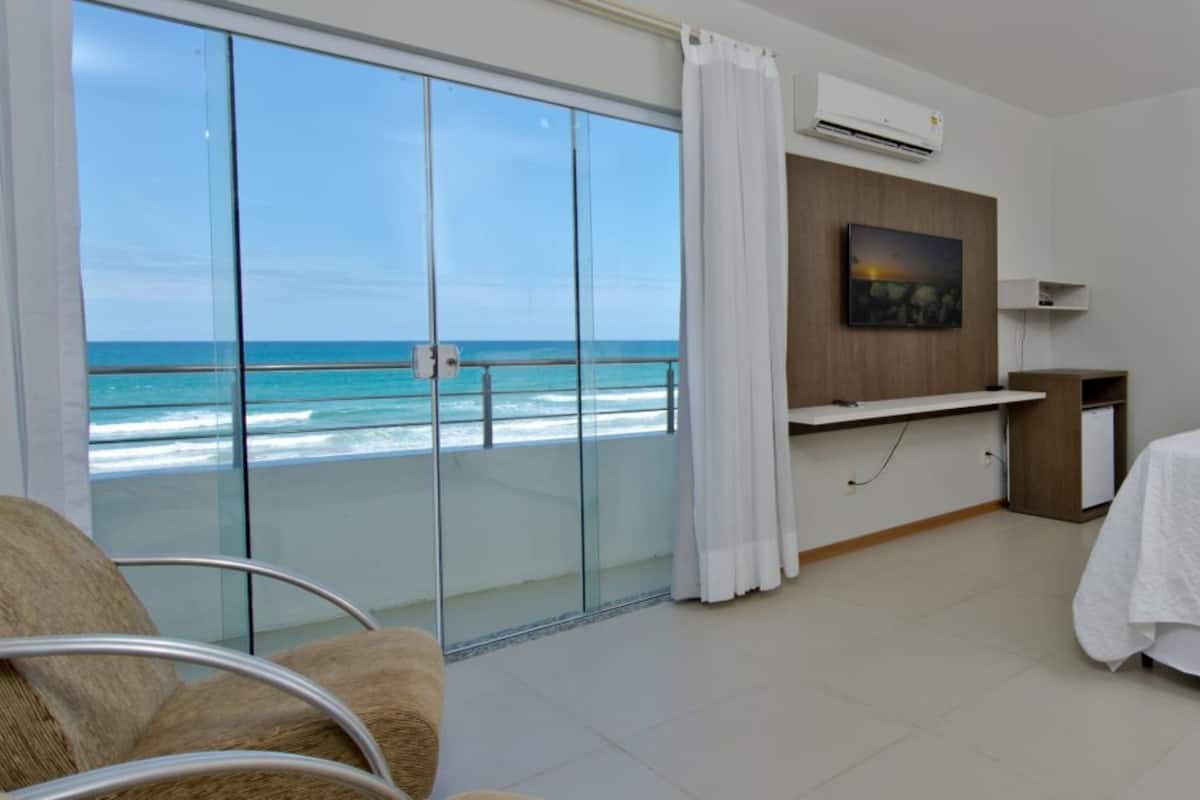 A hotel room with a bed, a chair, a desk, and a view of the ocean through large windows.