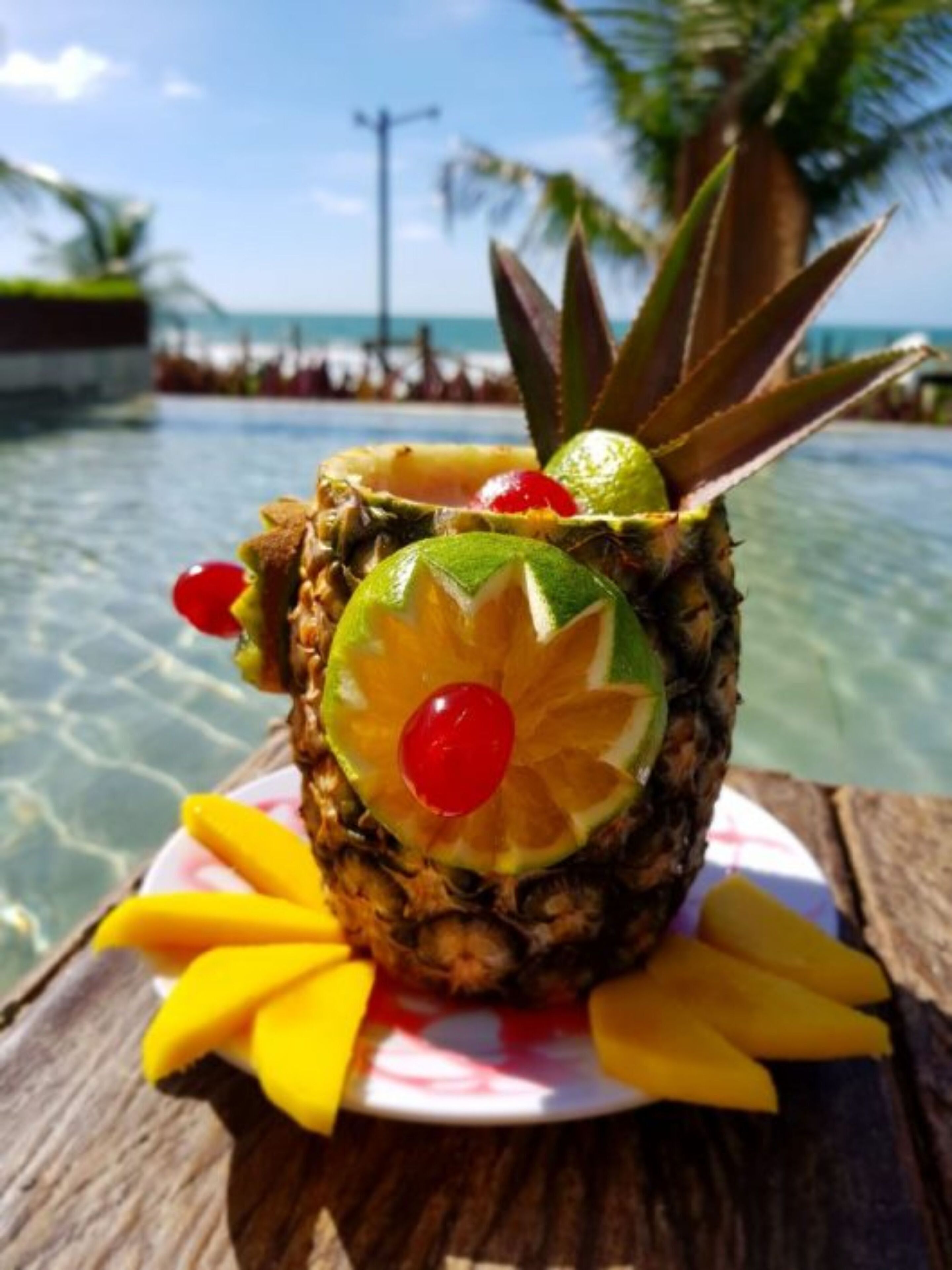 A pineapple with lime and cherry on a wooden table by a pool with clear water and palm trees in the background.