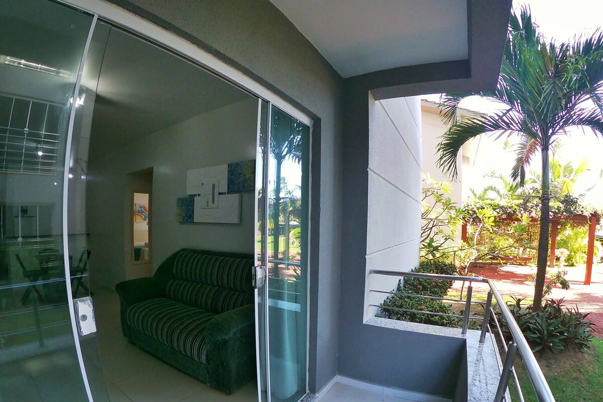 A modern living room with a green sofa, a glass door leading to a balcony, and a view of a garden with a palm tree.