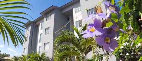 Un edificio moderno con amplias ventanas y balcones, rodeado de abundante vegetación y plantas en flor.