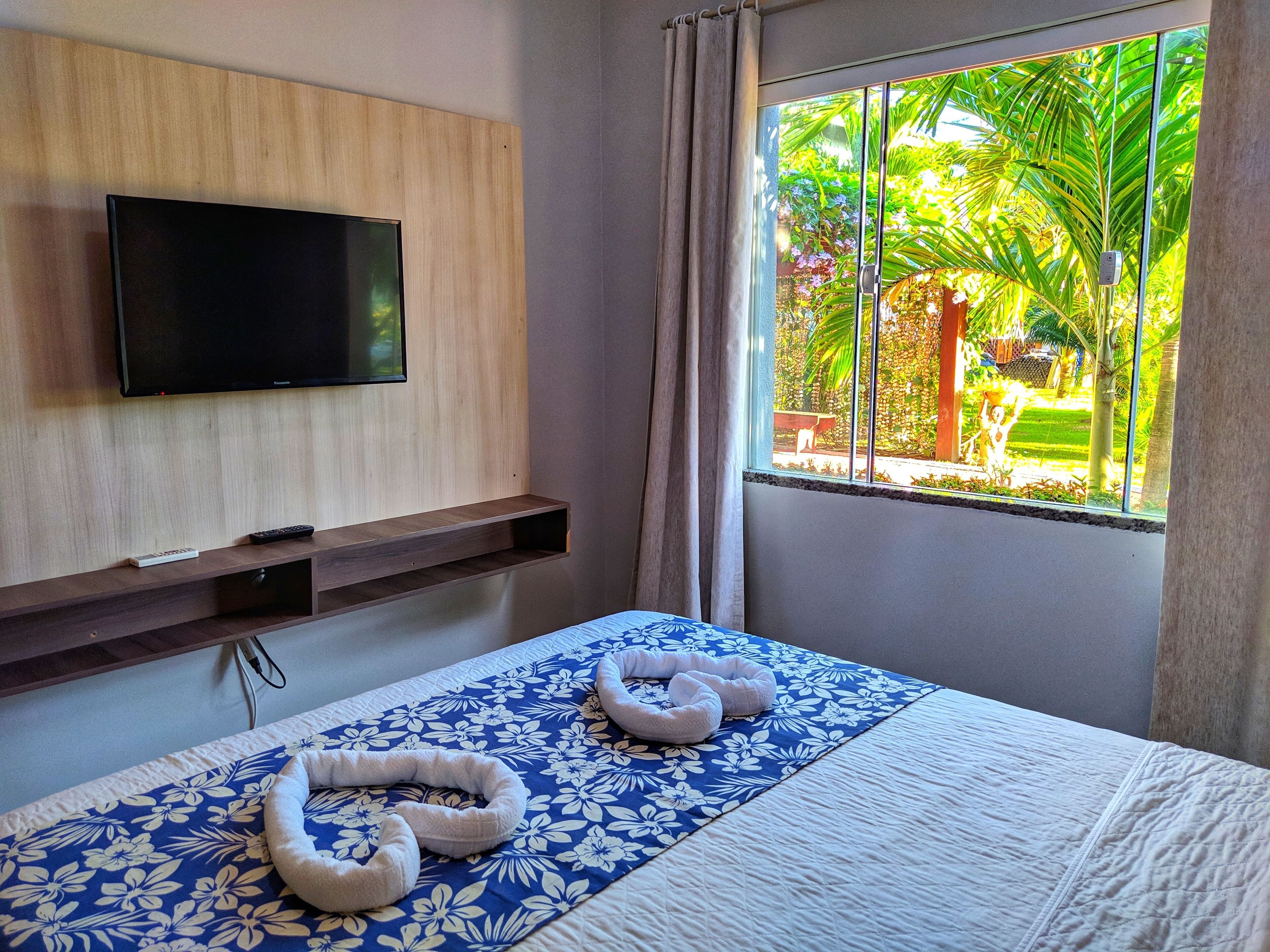 Una habitación de hotel con una cama, un televisor montado, una ventana con vistas a la vegetación y un estante de madera.