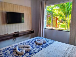 Una habitación de hotel con una cama, un televisor montado, una ventana con vistas a la vegetación y un estante de madera.