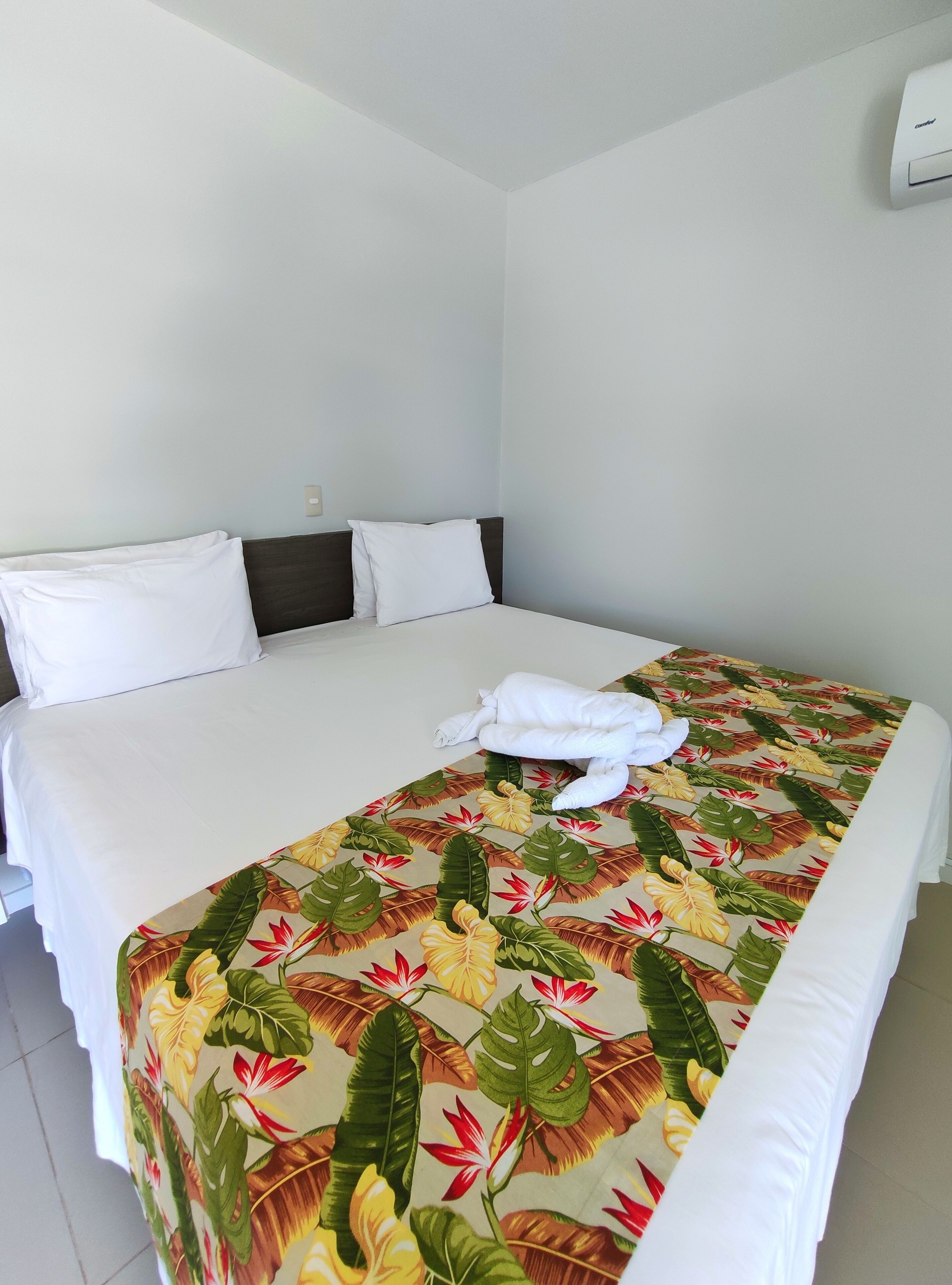 A hotel room with a bed, white bedding, a tropical leaf-patterned bedspread, and a white towel on the bed.