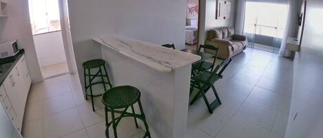 Una cocina moderna con encimera de mármol, dos taburetes verdes y una zona de comedor con sofá y sillas.