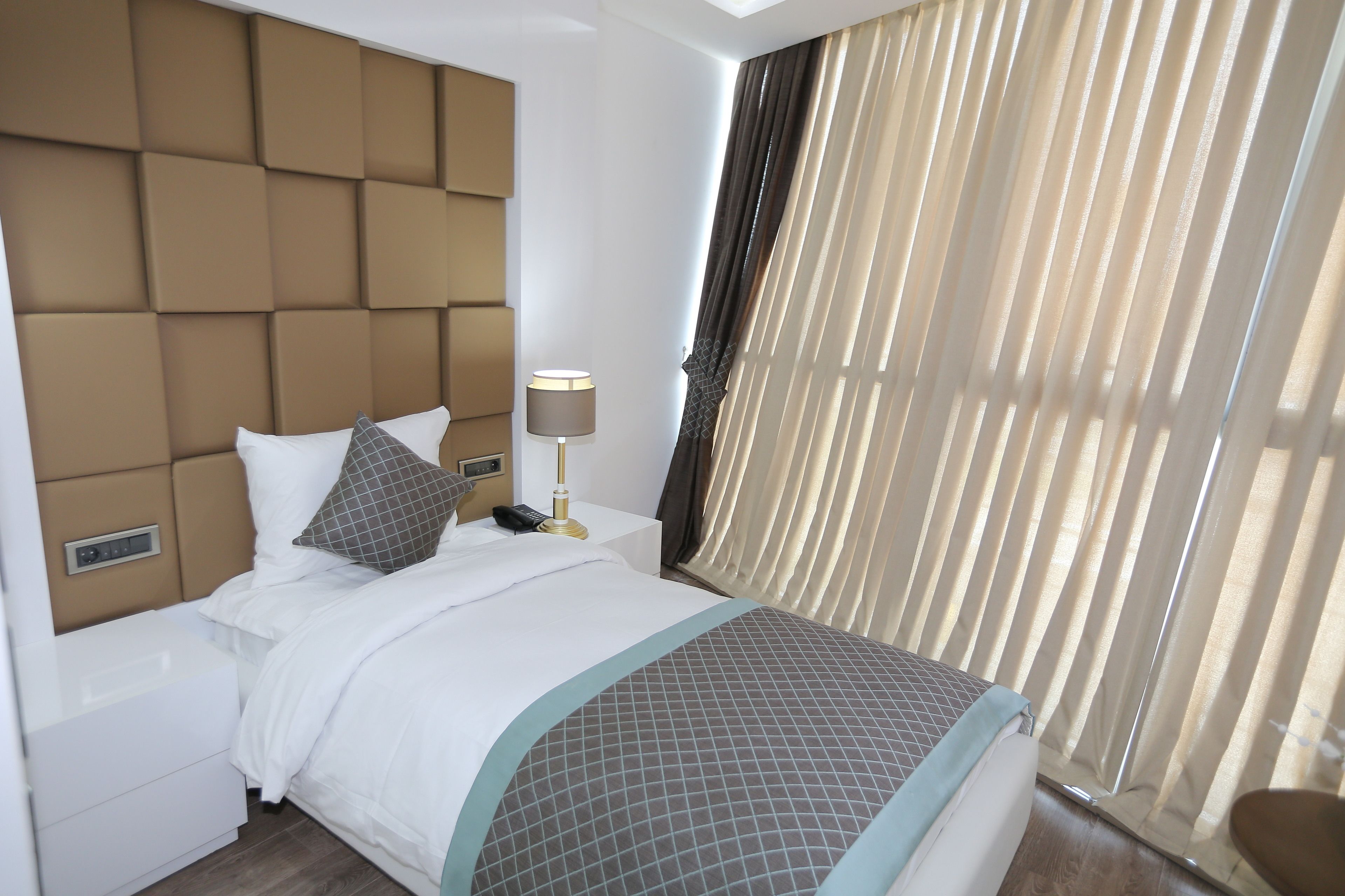 A hotel room with a large bed, a bedside table with a lamp, a control panel, and a window with curtains.